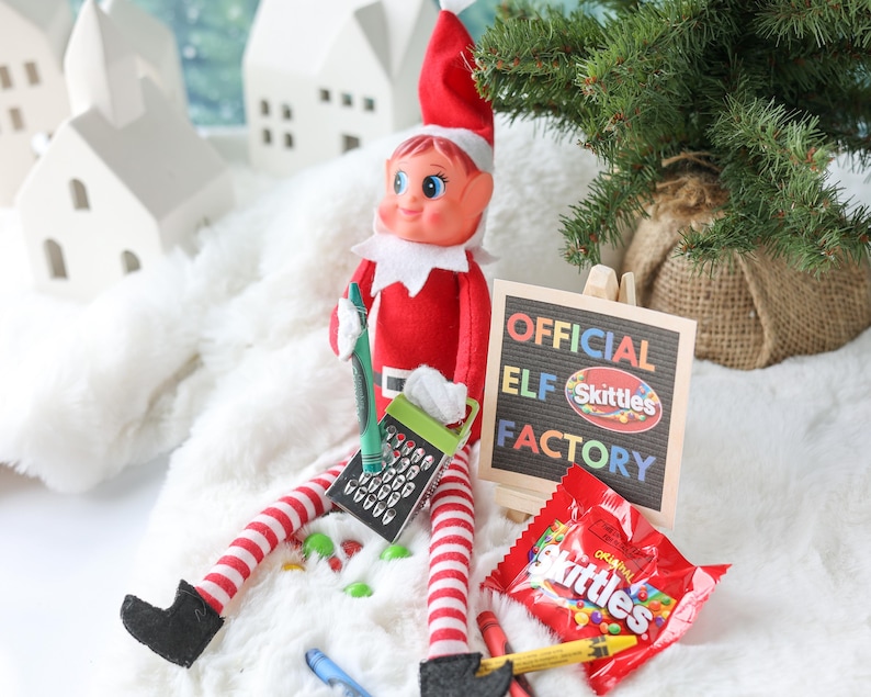 May include: A plush elf doll with a red suit and hat, red and white striped legs, and black boots. The elf holds a green crayon and a small metal grater. Next to it is a sign reading OFFICIAL ELF SKITTLES FACTORY and a bag of Skittles candy.