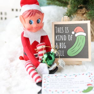 May include: A red and white elf doll holding a small green glass pickle ornament. Next to the doll is a wooden sign with the text "THIS IS KIND OF A BIG DILL" and a graphic of a pickle wearing a Santa hat. A small white card with text is also present.
