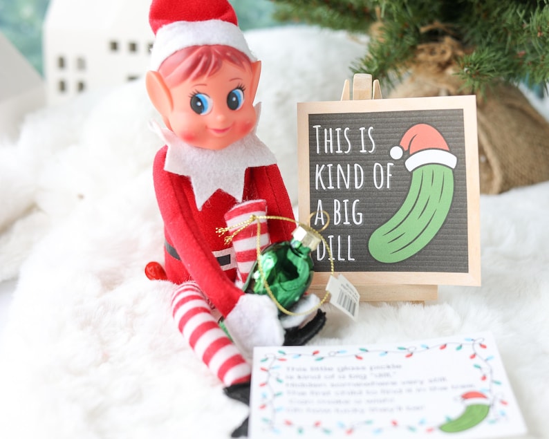 May include: A red and white elf doll holding a small green glass pickle ornament. Next to the doll is a wooden sign with the text "THIS IS KIND OF A BIG DILL" and a graphic of a pickle wearing a Santa hat. A small white card with text is also present.
