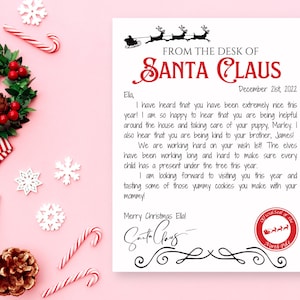 May include: A letter from Santa Claus on white paper with a pink background. The letter is addressed to "Ela" and includes a message, Santa's signature, and a North Pole seal. Christmas decorations surround the letter.