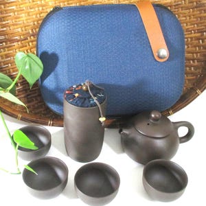 May include: A dark brown clay tea set consisting of a small teapot, four matching cups, and a tall cylindrical tea canister with a fabric lid. The set is accompanied by a blue textured carrying case with a tan leather strap and metal snap.