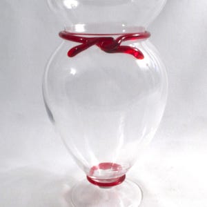 May include: A clear glass vase with a red glass ribbon around the middle. The vase has a round body and a flared top. The vase sits on a clear glass base.