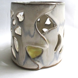 May include: A cylindrical ceramic candle holder with a white glaze and subtle blue-grey streaks. The exterior features large, irregular cut-out shapes that reveal a smooth interior surface. The base is unglazed, showing the natural brown clay.