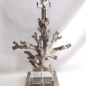 May include: A silver-toned table lamp featuring a coral-like design. The lamp has a textured, branching structure and sits atop a clear, square base. The lamp's metal stem supports a standard light bulb socket.