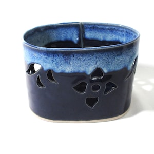May include: An oval ceramic vessel featuring a deep navy blue base color with a lighter blue glazed rim. The exterior wall is decorated with cut-out floral patterns, and the interior shows a smooth, dark blue finish with a visible vertical seam.