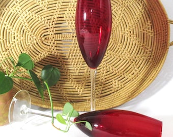 May include: Two tall champagne flutes with deep red glass bowls and clear glass stems. One flute stands upright while the other lies horizontally. The red glass has a subtle, swirling texture throughout the surface of the bowls.