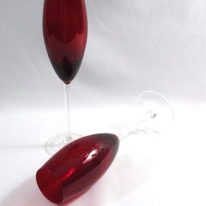 May include: Two elegant red wine glasses with long stems. One glass stands upright, while the other lies on its side. The glasses have a deep red color that fades to a darker shade at the base, creating a sophisticated look.