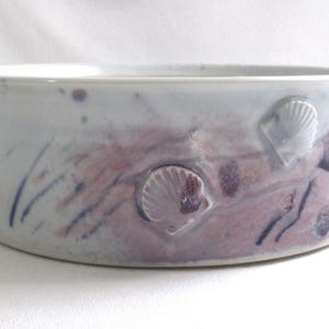 May include: A white ceramic bowl with a pink and blue glaze. The bowl has three raised seashells on the side and a scalloped rim.