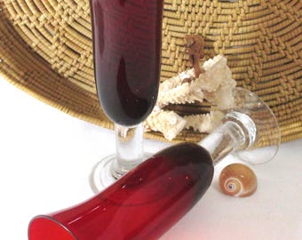 May include: Two tall, slender drinking glasses with deep red, trumpet-shaped bowls and clear glass stems and bases. One glass stands upright while the other rests horizontally, showcasing the vibrant red color and smooth, curved glass construction.
