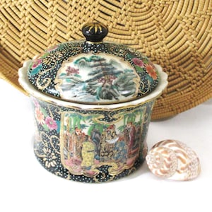 May include: A black ceramic lidded jar featuring intricate gold-trimmed panels with painted scenes of figures and landscapes, accented by colorful floral motifs and a black knob handle, accompanied by a small, patterned spiral seashell.