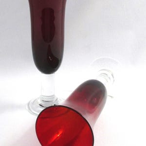 May include: Two tall, elegant wine glasses with a deep red hue. One glass stands upright, while the other lies on its side. The glasses have a clear stem and base, creating a striking contrast with the colored bowl.