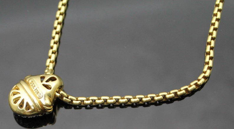 May include: A gold-colored necklace with a unique pendant. The pendant has a rounded shape with cut-out designs and is connected to a box chain. The necklace is made of a shiny, metallic material.