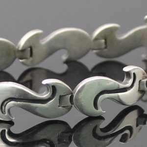 Op de afbeelding: Een zilveren armband met een golvend, abstract ontwerp. De armband is gemaakt van met elkaar verbonden, in elkaar grijpende stukken.