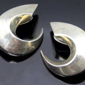 May include: A pair of silver hoop earrings with a curved, abstract design. The earrings are smooth and shiny.