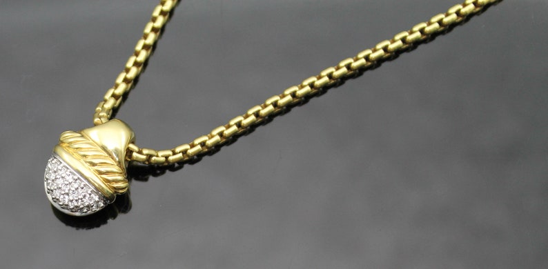 May include: A gold-toned necklace with a pendant featuring a cluster of small, sparkling stones. The pendant has a gold-colored top and a silver-colored bottom. The chain is a box chain style. The necklace is displayed on a dark surface.