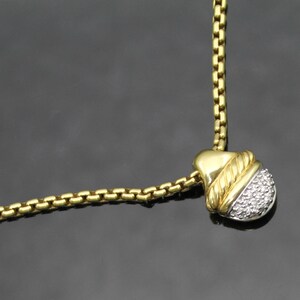 May include: A gold-toned necklace with a pendant featuring a gold-colored top section and a lower section encrusted with small, clear stones. The chain is a box-style design. The background is a dark gray.