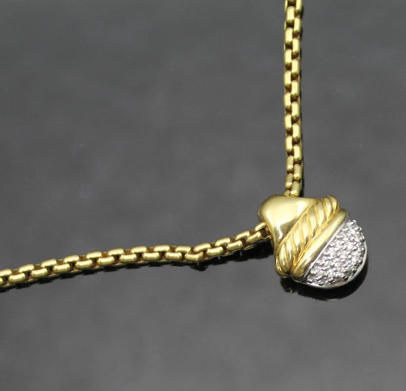 May include: A gold-toned necklace with a pendant featuring a gold-colored top section and a lower section encrusted with small, clear stones. The chain is a box-style design. The background is a dark gray.
