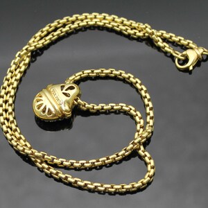 May include: A gold-colored necklace with a pendant. The necklace chain is made of small, square links. The pendant is a stylized shape with cut-out details. The clasp is also gold-colored.