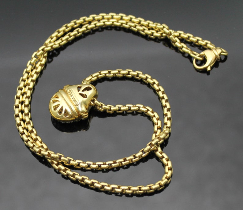May include: A gold-colored necklace with a pendant. The necklace chain is made of small, square links. The pendant is a stylized shape with cut-out details. The clasp is also gold-colored.