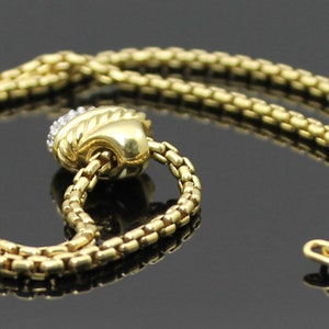 May include: A gold-toned necklace with a textured chain design. The necklace features a decorative pendant with a diamond-like embellishment. The clasp is visible at the end of the chain. The jewelry is displayed on a reflective surface.