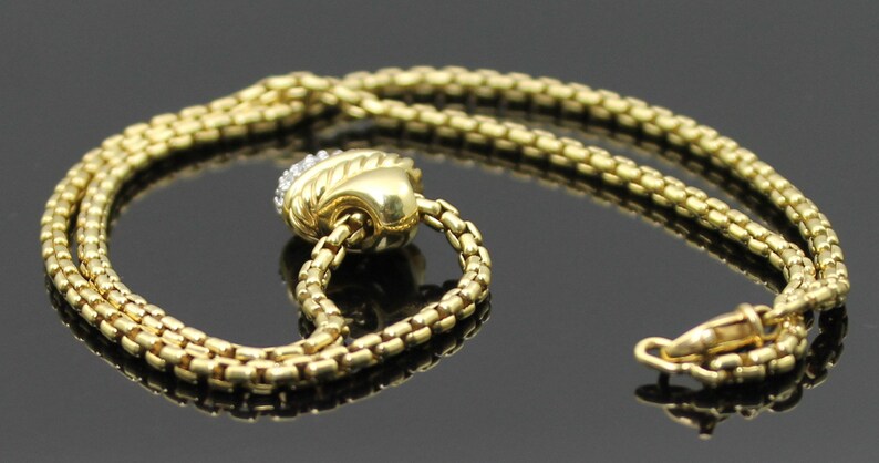 May include: A gold-toned necklace with a textured chain design. The necklace features a decorative pendant with a diamond-like embellishment. The clasp is visible at the end of the chain. The jewelry is displayed on a reflective surface.