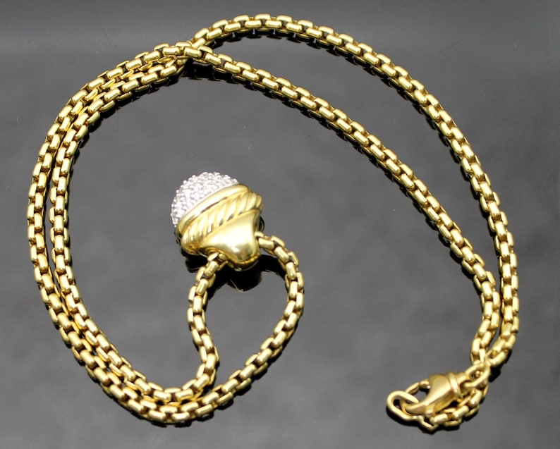 May include: A gold-toned necklace with a round pendant encrusted with small, clear stones. The pendant is attached to a chain with a lobster clasp. The necklace is displayed on a dark surface.