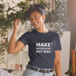 Peut inclure: T-shirt bleu marine avec l'inscription blanche « MAKE HAYSTACKS NOT WAR » et une illustration de fourchette. Le t-shirt est de style col rond.