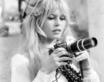 May include: A monochrome image of a woman with blonde hair holding a vintage camera. She wears a white blouse and a black and white polka dot accessory. The camera has a long lens and a black strap. The photograph has a classic, timeless feel.