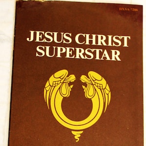 Könnte beinhalten: Ein braunes Programmheft für die Rockoper "Jesus Christ Superstar" von Andrew Lloyd Webber und Tim Rice. Das Cover zeigt eine goldene Illustration von zwei Engeln mit Flügeln und einem Heiligenschein, die einen Kreis mit einem stilisierten Kreuz umgeben. Der Text "Oktober 1970" ist unter der Illustration gedruckt.