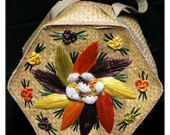 Könnte beinhalten: Eine sechseckige geflochtene Strohtasche mit einem braunen Ledergriff. Die Tasche ist mit einem großen Blumendesign aus buntem geflochtenem Stroh verziert. Die Blume hat eine Mitte aus weißen Muscheln und ist von orangefarbenen, gelben und braunen Blütenblättern umgeben. Die Tasche trägt den Schriftzug "Tessie" auf der Unterseite.