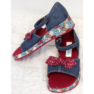 May include: A pair of blue denim sandals with red and white patterned fabric trim. The sandals have a bow on the front and a strap around the ankle. The sandals are on a white surface.