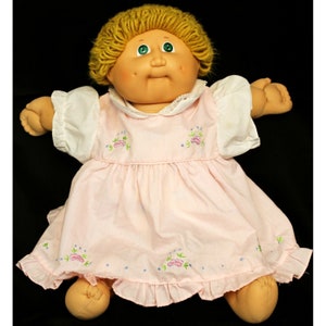 May include: A Cabbage Patch Kid doll with blonde yarn hair, green eyes, and rosy cheeks. The doll is wearing a pink dress with floral embroidery and a white lace collar.