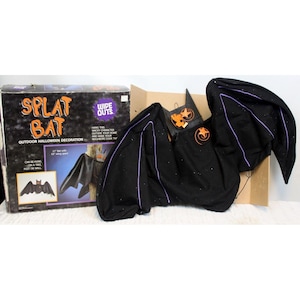 May include: A black outdoor Halloween decoration in the shape of a bat with purple accents. The bat has a 31-inch wingspan and is displayed with its original box. The box reads "Splat Bat" and "Wipe Outs."