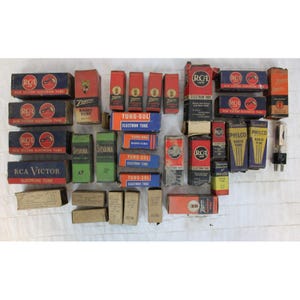 May include: A collection of vintage radio tubes in various brands and colours, including RCA Victor, Zenith, Sylvania, Tung-Sol, and Philco. The tubes are arranged in rows on a white surface.