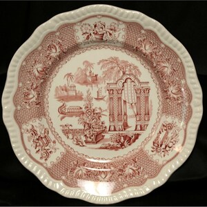 Może przedstawiać: Biały ceramiczny talerz z falistym brzegiem i szczegółowym czerwono-białym wzorem. Talerz przedstawia centralną scenę krajobrazową z budynkami, drzewami i łodzią, otoczoną kwiatowymi i geometrycznymi wzorami. Talerz jest elementem dekoracyjnym.