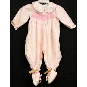 May include: A pink baby onesie with a white collar and pink and white embroidered details. The onesie has a pink and white pattern and has attached booties.