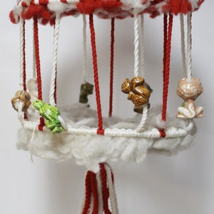 May include: A red and white striped hanging mobile with a white fluffy base and ceramic animal figurines. The mobile has a red and white tassel hanging from the bottom.
