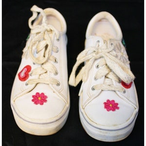 May include: A pair of white canvas sneakers with pink and red embroidered hearts and flowers. The laces are white and the shoes have a white rubber sole.