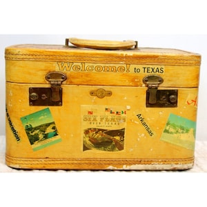 Könnte beinhalten: Ein vintage gelber Koffer mit einem braunen Ledergriff und einem "Willkommen in Texas" Aufkleber. Der Koffer hat mehrere andere Aufkleber, darunter einen für Six Flags Over Texas und einen für Arkansas.