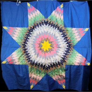 May include: A large blue quilt with a star-shaped pattern in the center. The star is made up of various colored fabric pieces, including pink, green, yellow, and white. The quilt is a traditional patchwork design.