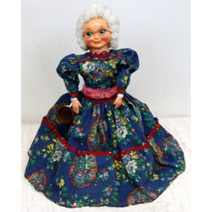 Può includere: Una bambola vintage con capelli bianchi, che indossa occhiali e un vestito blu a fiori con una cintura rosa. La bambola tiene una tazza marrone.