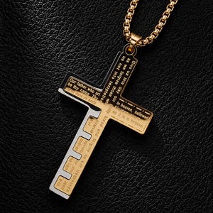 May include: A gold-toned metal cross pendant on a matching box chain. The cross features a two-tone design with black recessed areas containing engraved text from the Lord's Prayer, including phrases like Our father, who art in heaven.