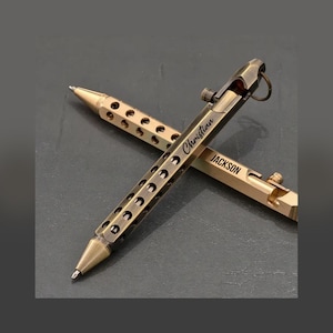 May include: Two bronze-colored pens are displayed against a dark background. One pen is engraved with the name "Christian," and the other with "JACKSON." Both pens have a series of circular cutouts along the barrel and a clip.
