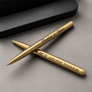 Op de afbeelding: Twee goudkleurige metalen balpennen die over elkaar liggen. Eén pen is gegraveerd met de tekst Madeline & Joshua in een schrijflettertype. De andere pen bevat turfstreepjes en de tekst years down, forever to go in het zwart.