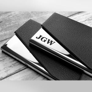 May include: A rectangular business card holder featuring a polished silver metal finish and a black textured leather cover. The silver metal surface is engraved with the letters JGW in a bold, serif font. The item is shown in a high-contrast black and white.