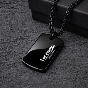 May include: Black dog tag pendant necklace with the text "BE STRONG JOSHUA 1:9" in white. The pendant is rectangular with rounded corners and hangs from a black chain. The necklace is displayed on a dark gray textured surface.