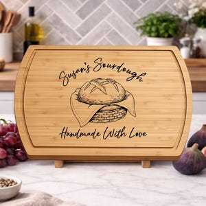 May include: A rectangular bamboo cutting board with rounded edges. The board features the text "Susan's Sourdough" and "Handmade With Love" along with a graphic of a loaf of bread in a basket. The board is supported by two small wooden feet.