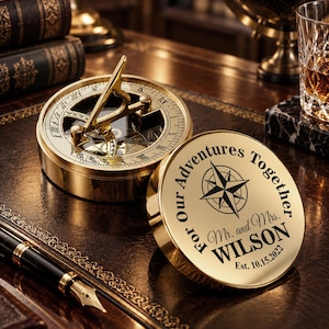 Könnte beinhalten: Ein polierter Messing-Sonnenuhr-Kompass mit klappbarem Gnomon und kreisförmigem Deckel. Der Deckel ist mit einer Windrose und dem Text graviert: For Our Adventures Together, Mr. and Mrs. WILSON, Est. 10.15.2022. Das Gerät zeigt römische Ziffern.