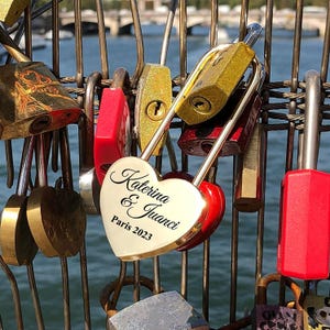 May include: A collection of love locks, including a heart-shaped lock with the names "Katerina & Juanci" and "Paris 2023" engraved on it. The locks are in various colors, including gold and red, attached to a metal structure.