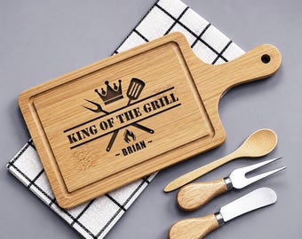 May include: A wooden cutting board with a handle, engraved with "KING OF THE GRILL" and a name. Includes a wooden spoon, fork, and knife. The board rests on a checkered cloth, all on a gray surface.